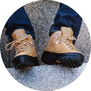 Image of a pair of Stüssy X Timberland brown boots which contain a black sole paired with dark denim jeans. The boots are facing forward towards the camera and the image is from the model's mid-shin downwards with the boots resting on a granite stair.