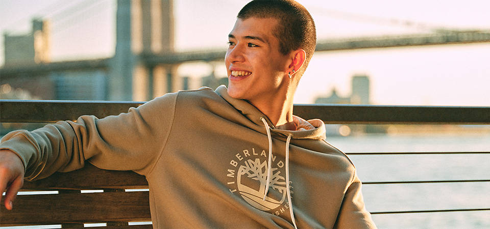 Image of a man with close-cropped hair, wearing a brown Timberland hoodie with a tan Timberland tree logo on the front, smiling and sitting on a bench by a river, with a bridge overhead.