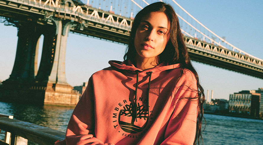 Image of a woman with long loose dark hair, wearing a red Timberland logo hoodie, leaning against a railing beneath a large suspension bridge under a blue sky.