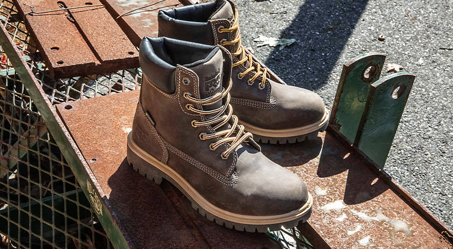 Image of a closeup of two brown work boots with black collars, sitting on some planks and metal grates outside.