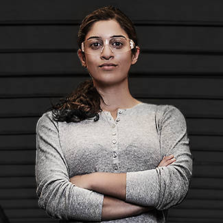 Image of a woman from the waist up, with brown hair in a pony tail standing in front of a black garage door. She is wearing safety glasses and a silver long sleeve shirt while her arms are crossed across her torso.