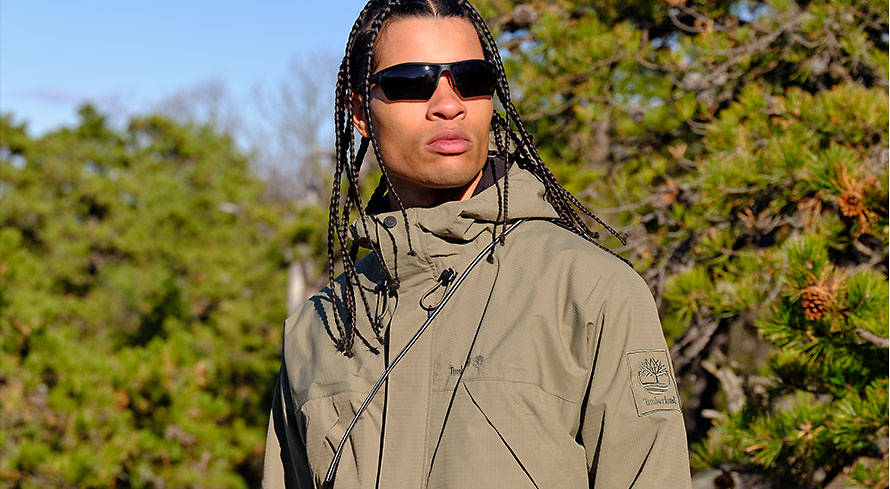 Image of a man in sunglasses and long braids, wearing a taupe colored jacket and standing with a backdrop of pine trees.