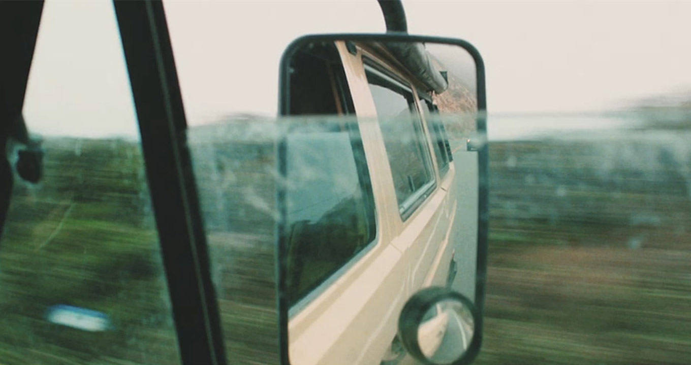 Image of the rear view mirror from a 1970s model van, driving along a mountainous road during the day with a mountainous terrain being reflected in the mirror.