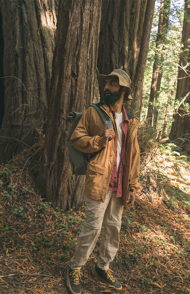 Image of a bearded man in a bucket hat, standing among trees in the woods in Timberland clothing and backpack, with black and wheat hiking boots.