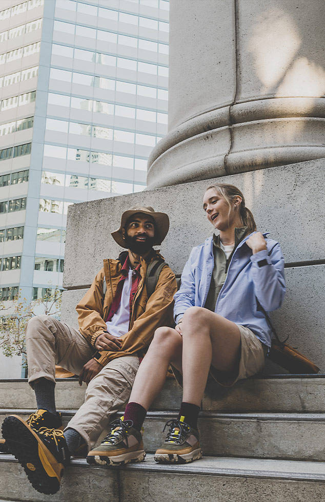 Image of a man and woman sitting on city steps against a large concrete pillar, wearing Timberland wheat and black hikers.