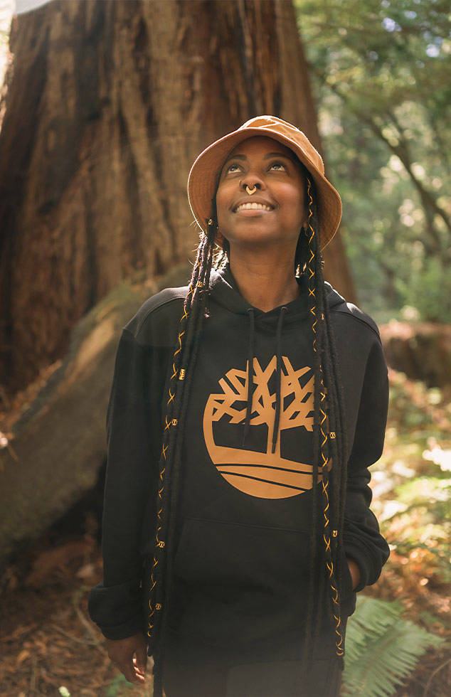 Image of a woman with long braids, bucket hat and Timberland hoodie, standing in the woods and looking up at the forest canopy.