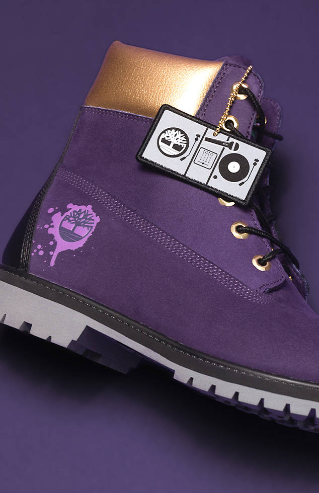 Overhead image of the Timberland Men's Hip Hop Royalty 6-inch Waterproof Boot on a purple background. The boot is purple with a lighter purple shade graffiti paint drip overlayed on top of the Timberland logo on the side of the boot. The ankle portion of the boot is gold while the eyelets are gold as well. The laces are black and the sole is gray. Includes a cloth badoink that looks like a DJ set up including the Timberland logo representing one of the records on the turntable.