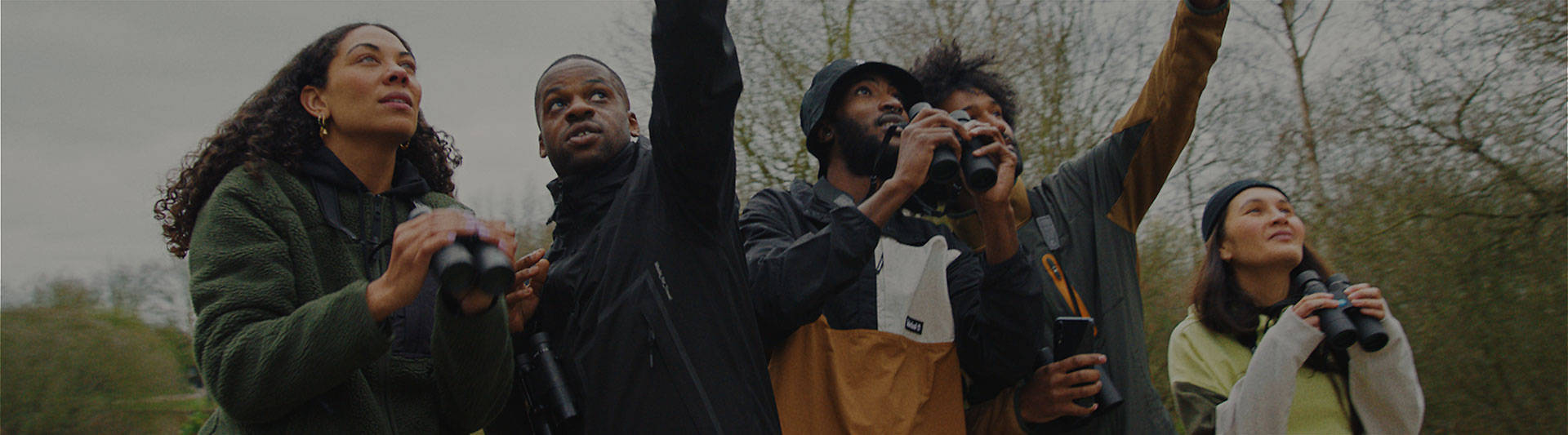 Image of five friends from the waist up, wearing Timberland gear on a cloudy day, standing out in a field with trees behind them, all holding binoculars and pointing up into the sky at something out of frame.