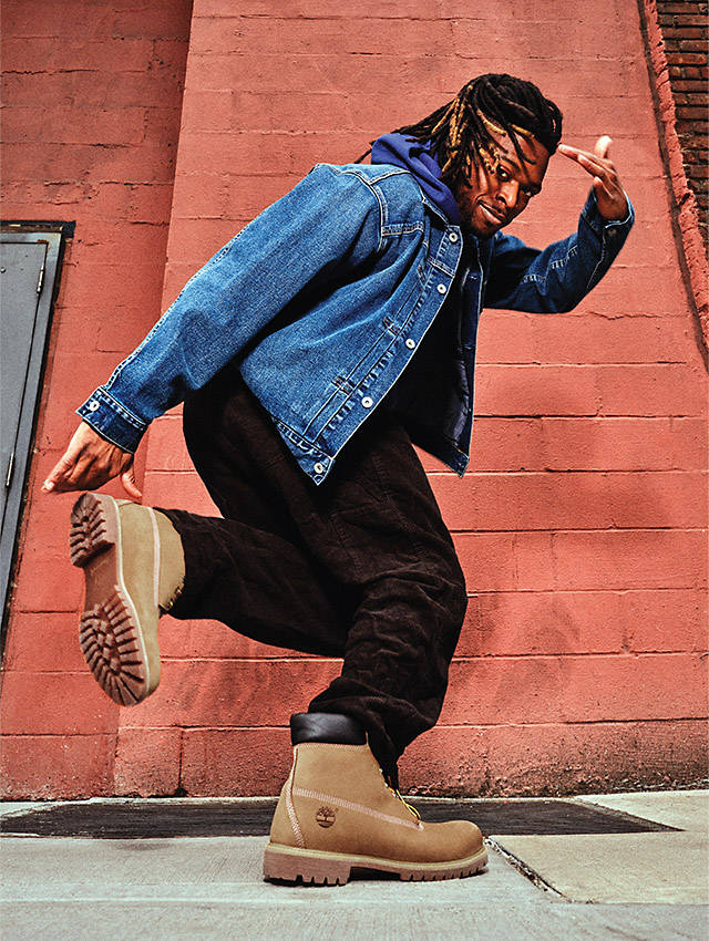 Image of a man with braids, wearing a blue jean jacket, dark pants and Timberland® classic boots, standing on one leg on a concrete alleyway against a red brick wall and saluting the camera.