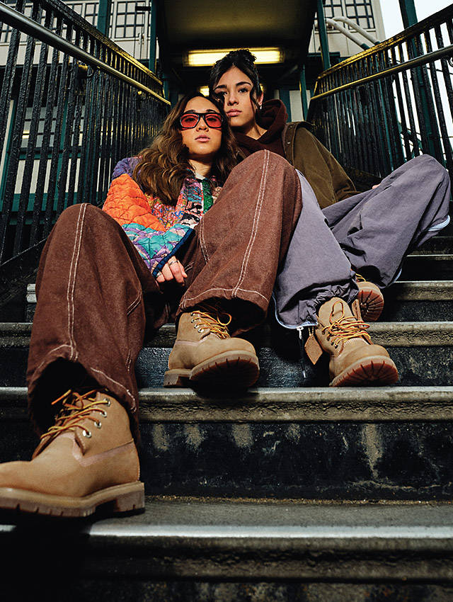 Image of two dark-haired young women sitting close together on a city stoop, one with orange-lensed sunglasses, with the camera angle putting the classic Timberland 6-inch boots they’re both wearing in the forefront.