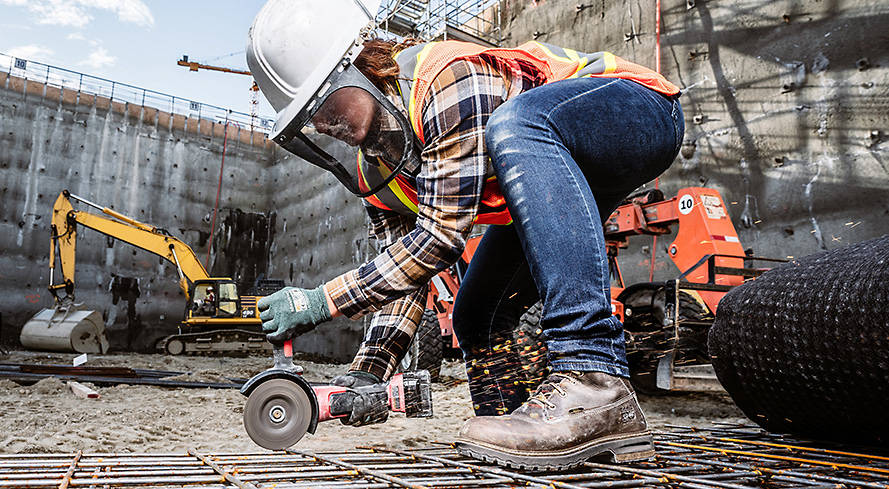 Image of a woman crouched with a tool in a construction zone, wearing a white hard hat with face shield, jeans, work flannel shirt and orange safety vest, with brown Timberland work boots.
