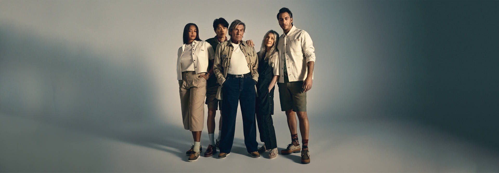 Image of five people of all ages and ethnicities, standing in muted clothing and Timberland boat shoes, facing the camera. Background is a blurred, darkened white room.