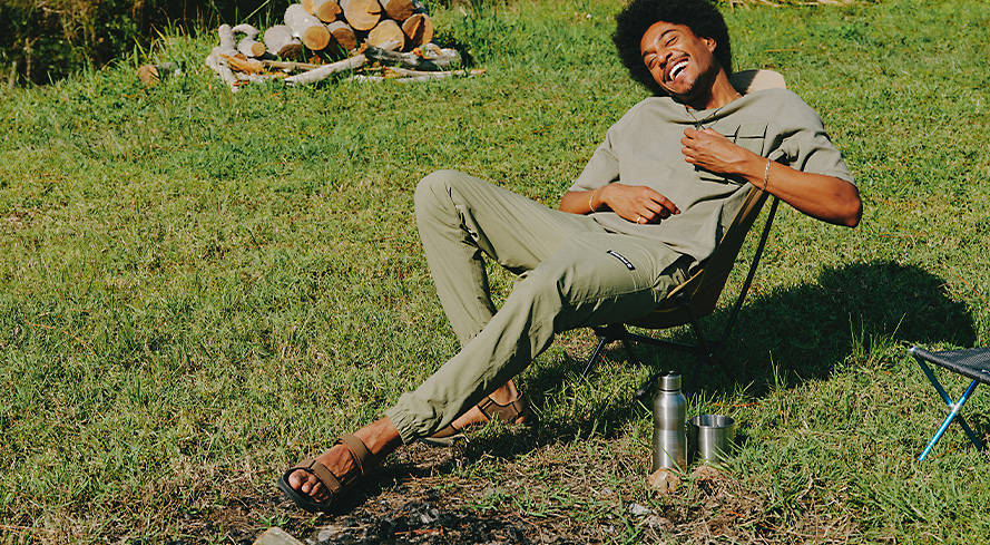 Image of a man in a lawn chair on grass, laughing in the sun, water bottle next to him, wearing a gray Timberland outfit and brown Timberland sandals.