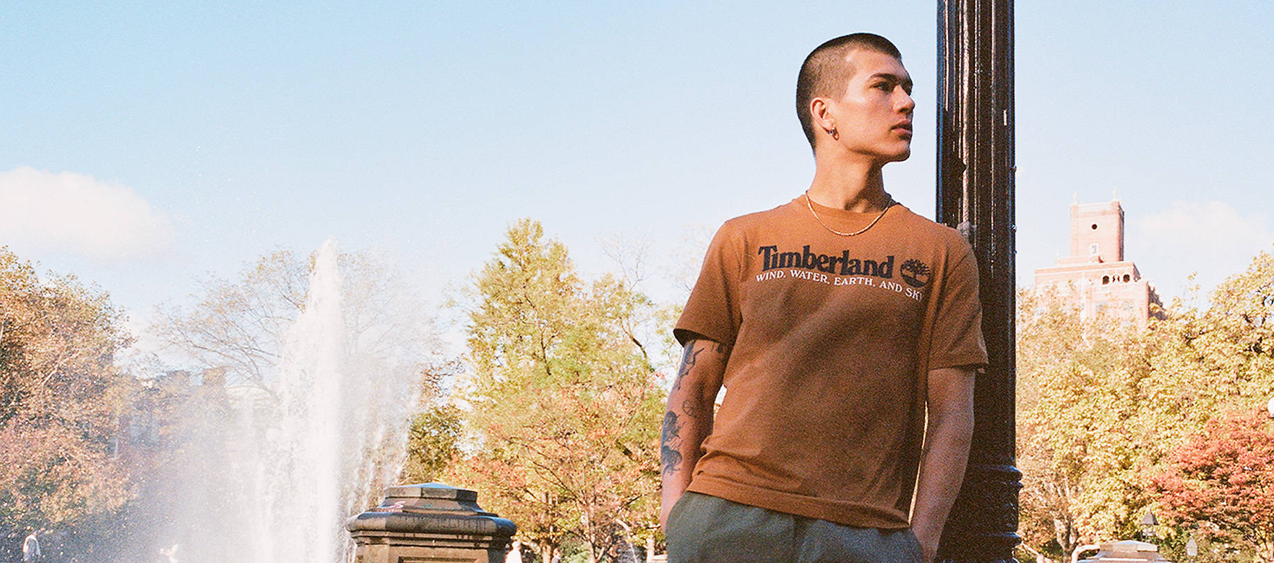 Image of a man in a forested city leaning against a lamp post, against a collage of a brown slip-on shoe, a brown Timberland t-shirt and nylon jogger pants.