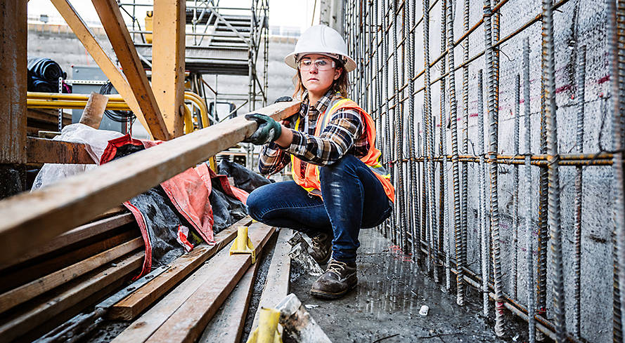 Image of a woman crouching in a manufacturing plant, wearing hard hat, orange vest, denim jeans and brown Hightower Timberland work boots.