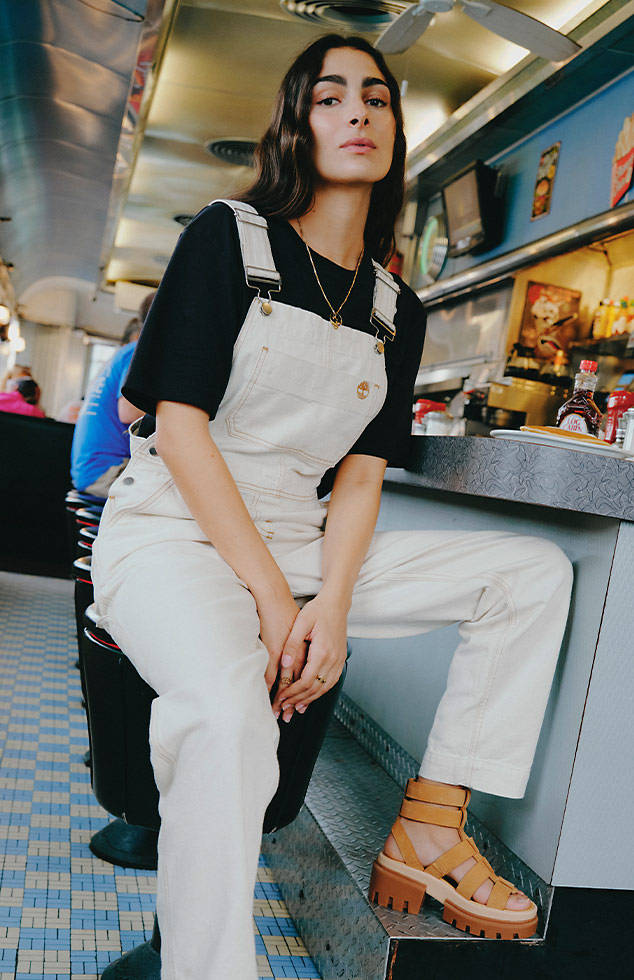 Photo of a woman in a black t-shirt, white Timberland overalls, and wheat-colored Timberland sandals seated at the counter of a diner.