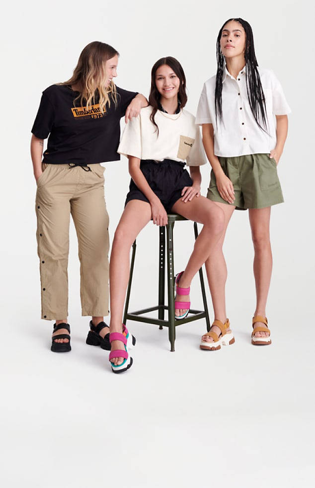 Image of three young women against a white background. One is wearing a black Timberland t-shirt, tan pants and black sandals. One is wearing a white tee and black shorts, with bright pink sandals. The third is wearing a white button-down shirt, green shorts and tan sandals.