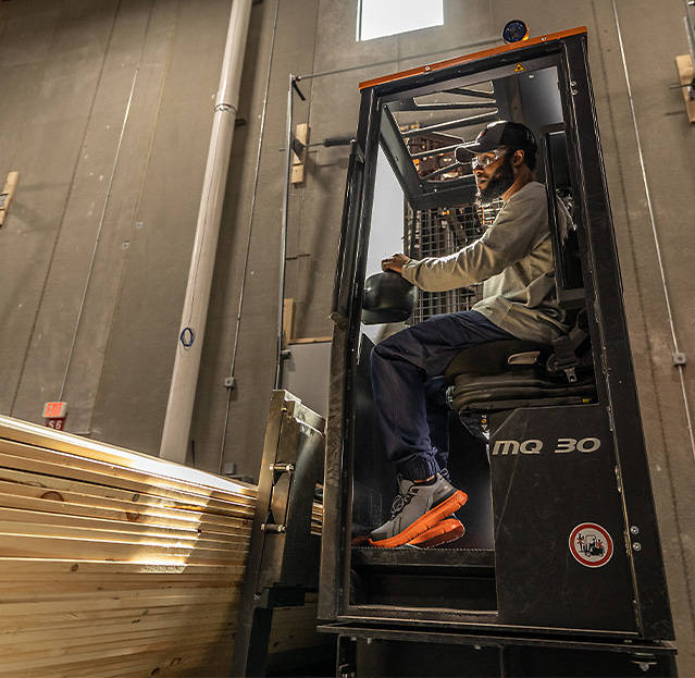 Image of a warehouse worker using heavy machinery, wearing a tan Timberland long sleeve shirt, dark blue Timberland jogger work pants and gray Timberland work sneakers with orange soles.