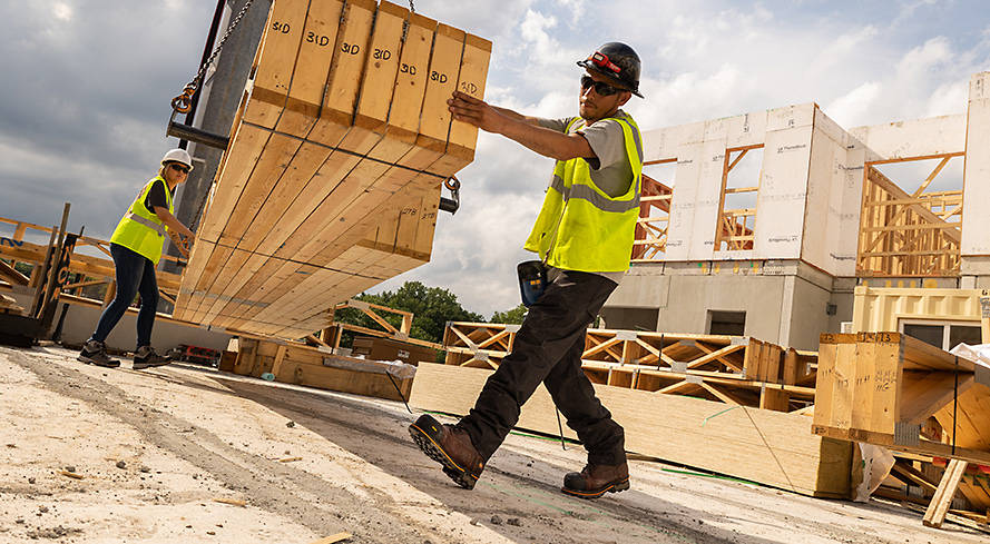 Image d'un homme sur un chantier de construction par temps nuageux, portant une chemise réfléchissante jaune, un pantalon de travail Timberland marron foncé et des bottes de travail Timberland marron, aidant à déplacer une palette de bois.