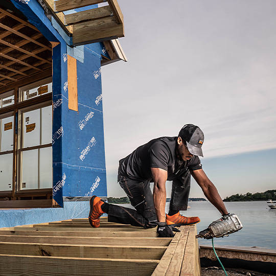 Image of a man in black Timberland work t-shirt and pants, wearing orange athletic work shoes, crouched on new construction with a nail gun against a backdrop of lake and sky.