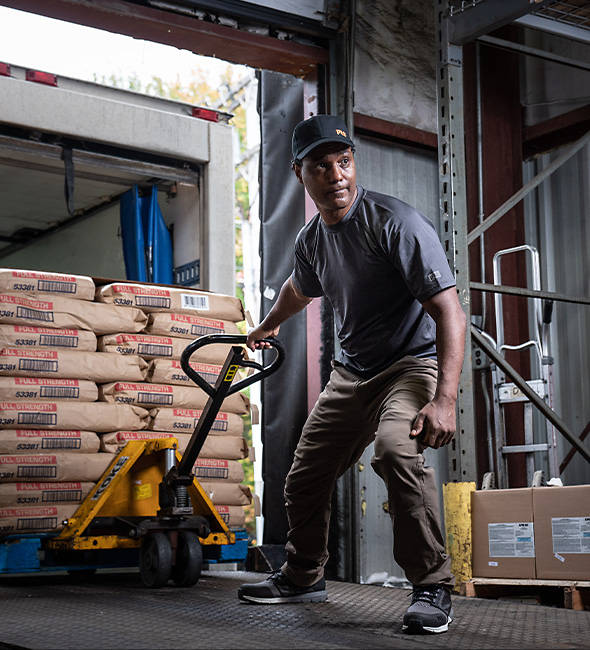 Image of a man in a warehouse, wearing dark Timberland work clothing and black work sneakers, pulling a pallet of concrete.