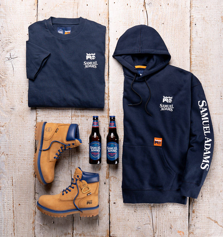 Overhead view of all Sam Adams x Timberland PRO products from this collaboration release on top of a wooden floor including: Beerproof T-Shirt, Beerproof Sweatshirt and Beerproof Boots. In the center of the picture, sandwiched between these products, are two bottles of Samuel Adams Lager.