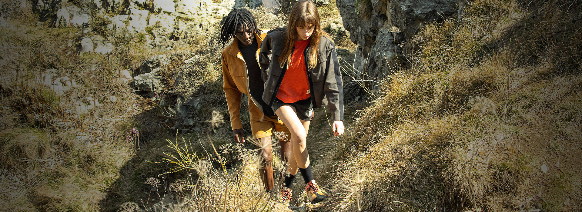 Image of two friends hiking toward the camera along a rocky mountain trail, wearing Icebreaker x Timberland brand wool jackets and chino shorts. The woman at front has long brown hair with bangs and an orange shirt; the man behind her has braids and is wearing a black shirt, and both are wearing hiking shoes.