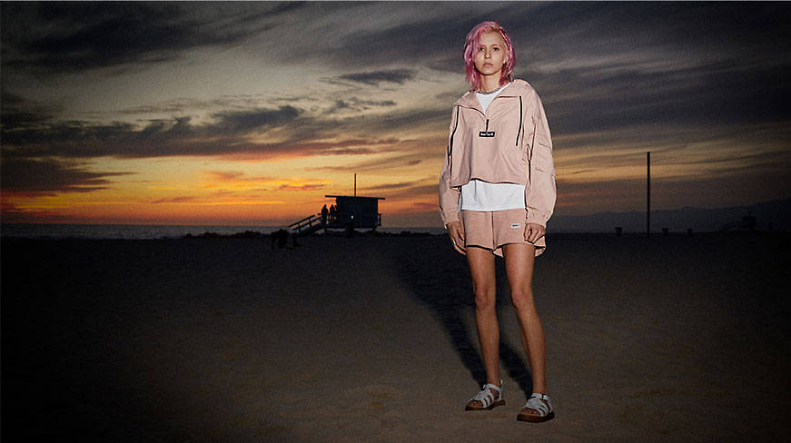Image of a woman in white strappy sandals, pink shorts and jacket, with pink hair, standing on a beach at sunset.