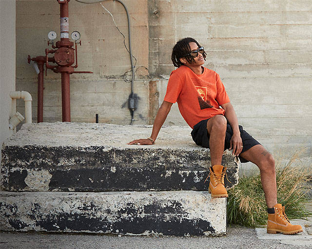Image of a man in classic wheat Timberland boots, orange tee, black shorts and sunglasses sitting on some concrete steps.