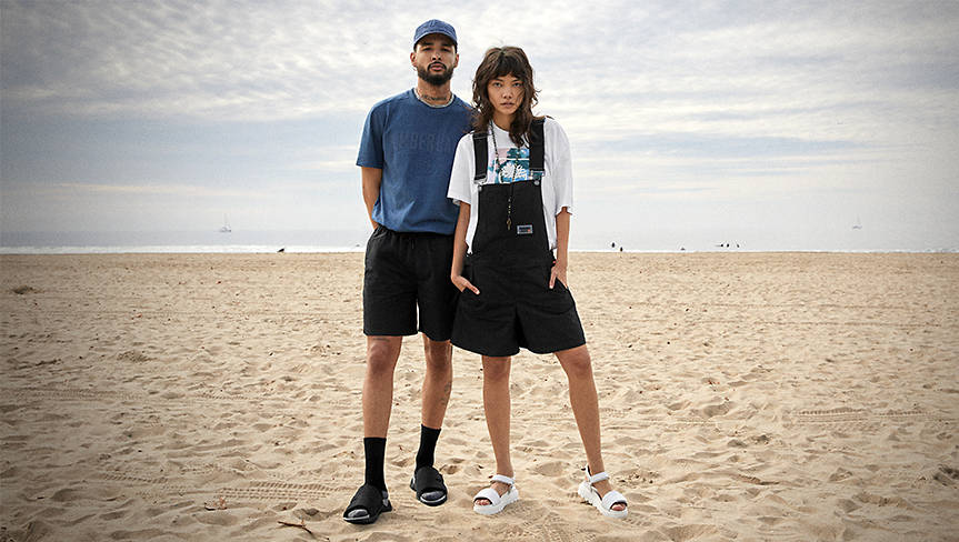Image of a man in black slide sandals and a woman in white strap sandals, standing together on a sandy beach.