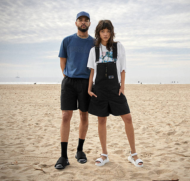 Image of man and woman on beach, him wearing black slide sandals with socks and she in white strappy sandals.