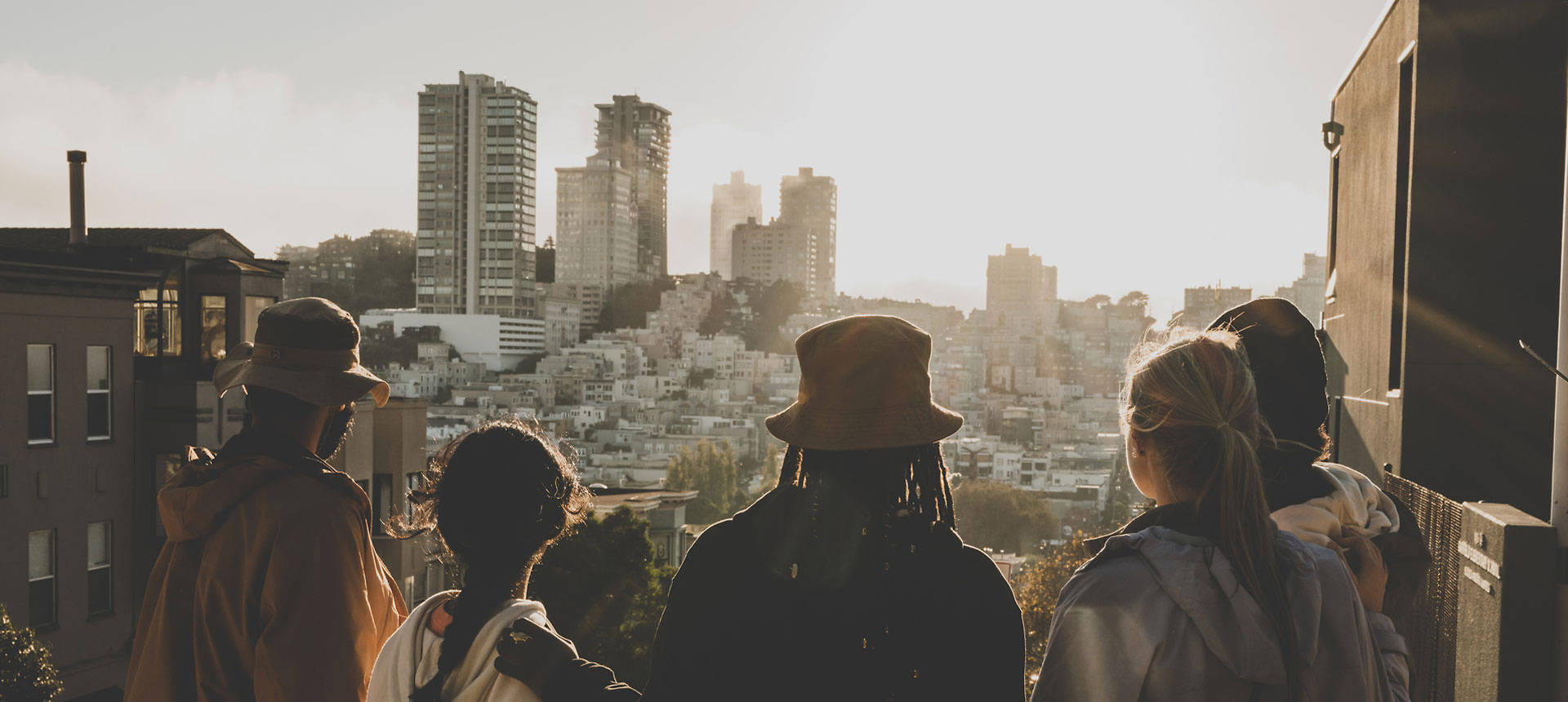 Image of five friends standing together and wearing Timberland jackets and hats, shown from the back. with the sun lighting up their silhouettes as they look out upon a city with white buildings.