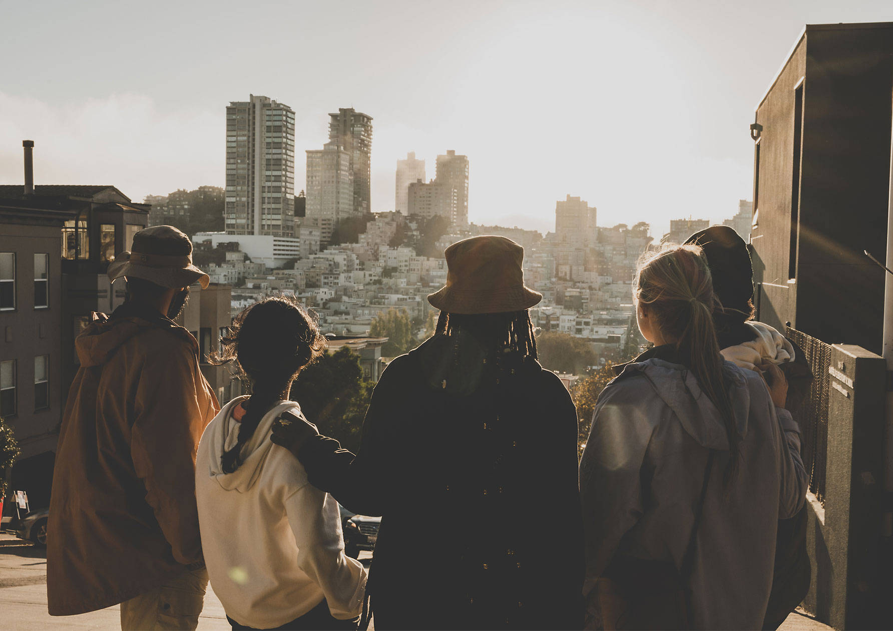 Image of five friends standing together and wearing Timberland jackets and hats, shown from the back. with the sun lighting up their silhouettes as they look out upon a city with white buildings.