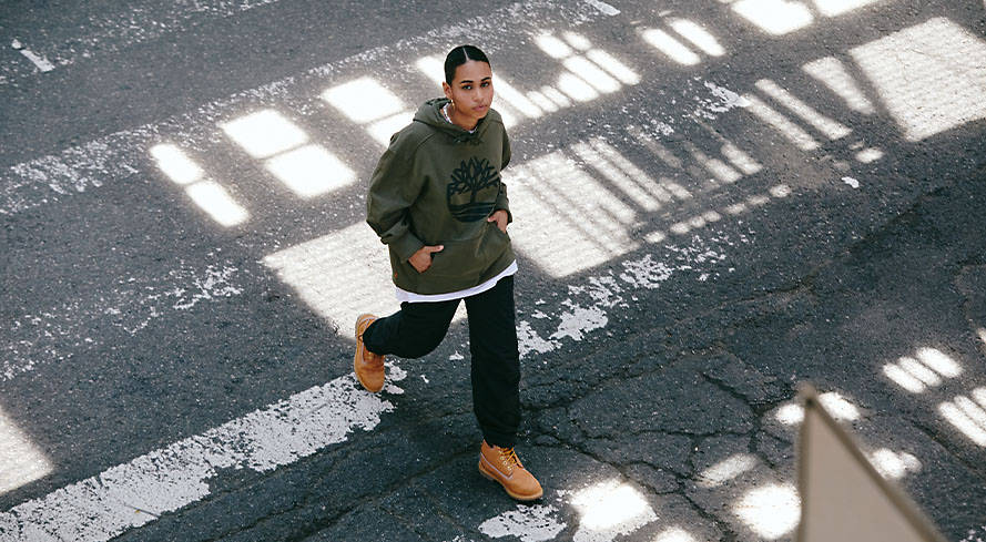 Image of a woman in a green Timberland tree logo hoodie, wheat boots and black pants walking across a city crosswalk.