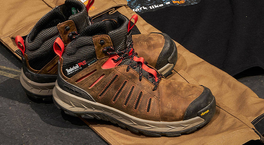 Close-up of a pair of two brown high-top hiking-style work boots, with pops of red color on a sheet of plywood.