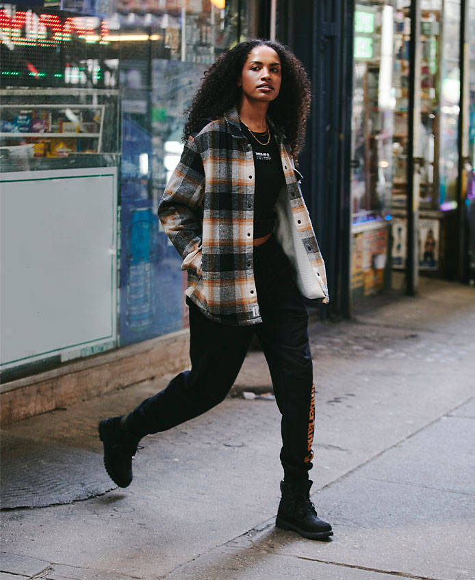 Image of a woman with long black curly hair, black and white flannel, black pants and black 6-inch Timberland boots crossing a street.
