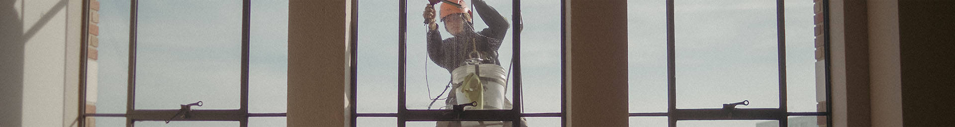 Image of a woman harnessed in the air, washing a window in boots and a hard had, with the image taken from inside the building so she's seen from the front.