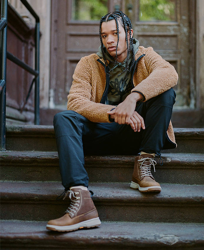 Image of a man in braids sitting on the stairs of a city stoop, wearing navy pants, a tan jacket and brown lace-up boots.