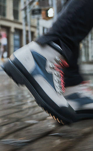 Blurry action image of a light gray Timberland™ boot with blue accents and red shoelaces, walking on a cobblestone sidewalk in the city.