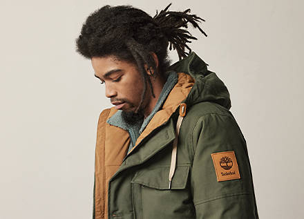Image of a man from the chest up, with dreadlocks, wearing an olive green jacket with brown leather accents, against a gray background.