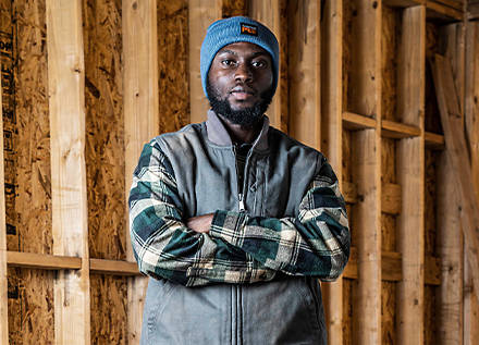 Image of a man in a blue winter beanie, flannel shirt and grey vest, against the unfinished framed wall of a new construction house.