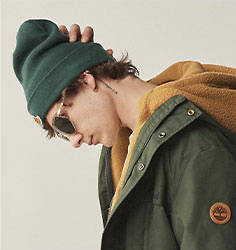 Image of a the side of the face of a young man with a green winter hat, green Timberland 3-in-1 winter coat and sunglasses. He is facing down with his right hand on the back of his head.