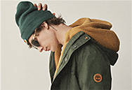 Image of a the side of the face of a young man with a green winter hat, green Timberland 3-in-1 winter coat and sunglasses. He is facing down with his right hand on the back of his head.