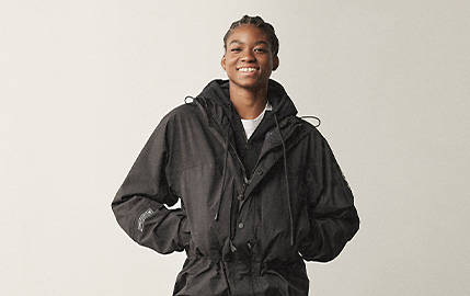 Image of a smiling woman in braids against a light grey background, with her hands in the pocket of a black jacket.