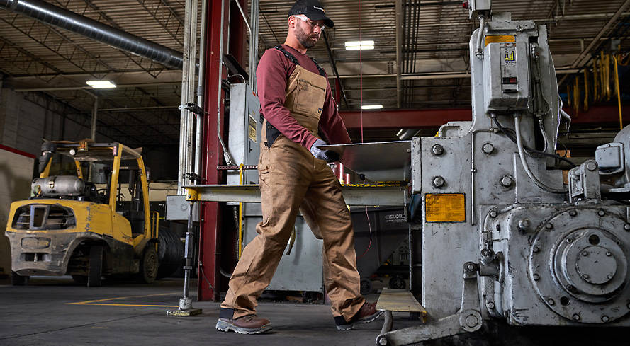 Image of a man in a warehouse next to some steel machinery, wearing tan overalls, red shirt, safety goggles and brown work boots with black outsoles.