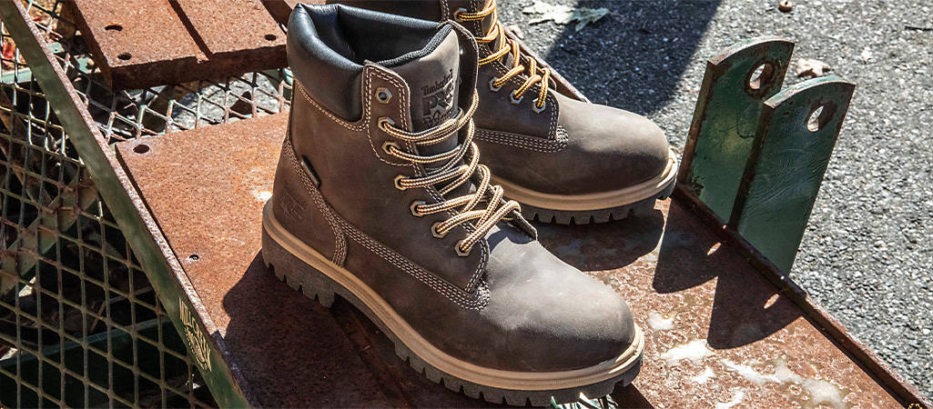 Image of a closeup of two brown work boots with black collars, sitting on some planks and metal grates outside. 