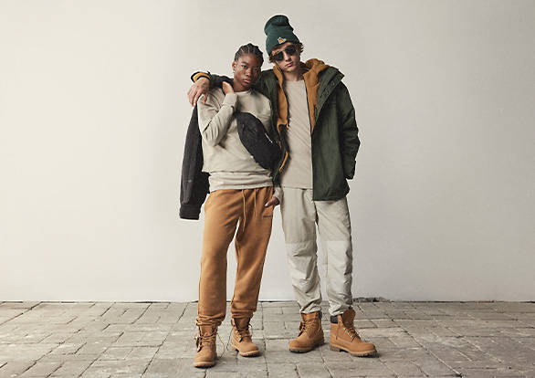 Image of a young woman and young man looking directly at the camera, standing on top of stone pavers. The man has his right arm over the shoulder of the woman and both are in a relaxed pose wearing Timberland winter gear including Timberland jackets, Timberland sweatpants and Timberland boots.