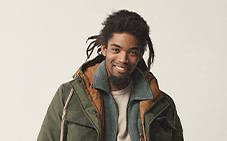 Image of a man from the chest up, with dreadlocks, smiling directly at the camera wearing an olive green Timberland 3-in-1 jacket with brown leather accents, against a tan background.