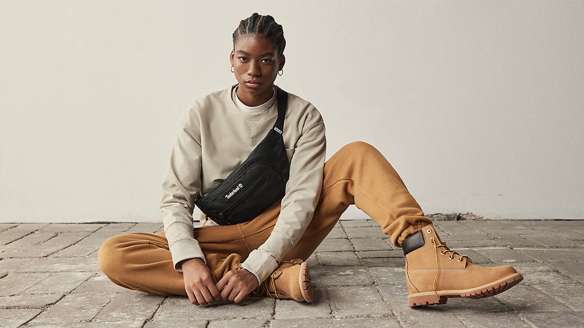 Image of a woman with braids wearing a black Timberland sling bag, being worn by an individual with an off-white long sleeve shirt, burnt orange sweatpants and Timberland boots.