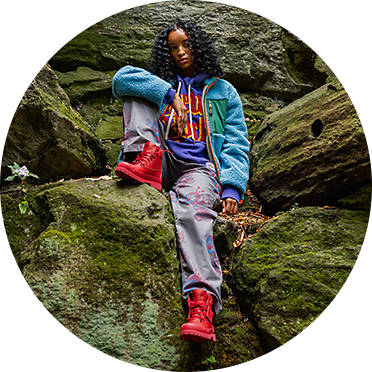 Image of a woman on mossy, large rocks in a forest looking down towards the bottom left while wearing Bee Line x Timberland clothing consisting of a blue jacket, light purple baggy pants and red Bee Line x Timberland boots.